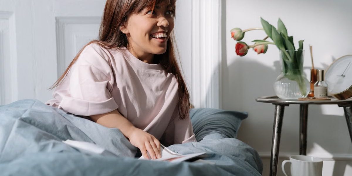 Asian woman with brown hair smiling, enjoying a calm morning in bed, surrounded by flowers and coffee.